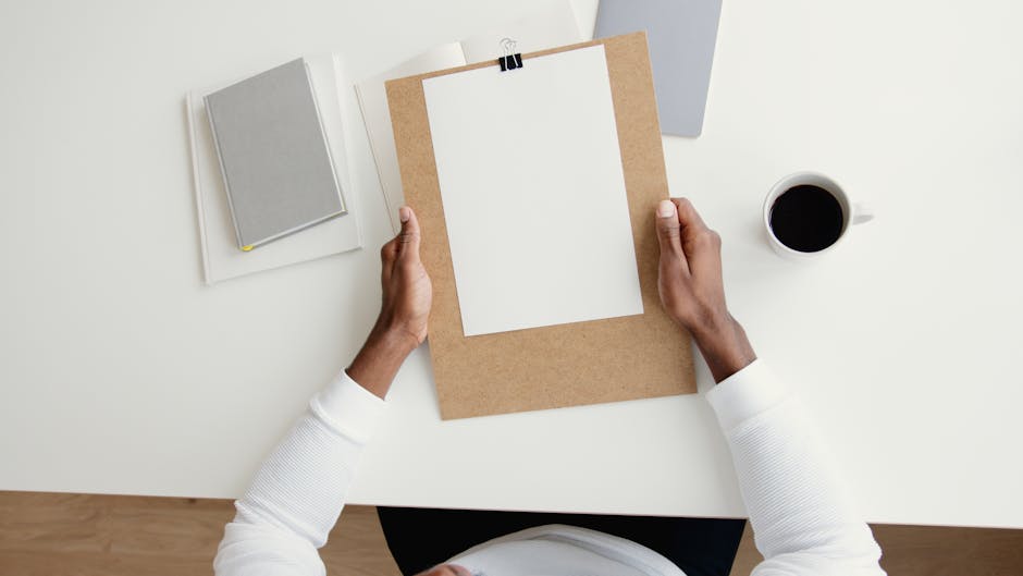 Top view of a person holding a clipboard with blank paper on a desk with coffee and books.