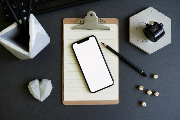 Flat lay of a smartphone on a clipboard with a pencil, ideal for tech mockups.