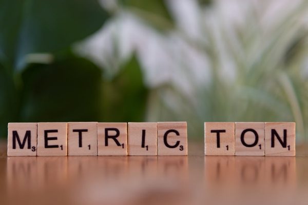 Scrabble tiles on a brown surface spell 'Metric Ton' with blurred green background.