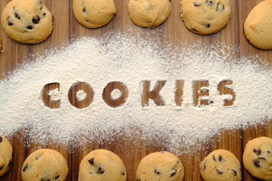 Freshly baked chocolate chip cookies arranged on flour-dusted wooden table.
