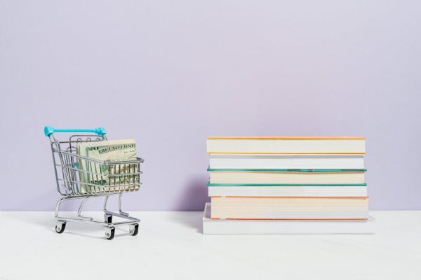 A shopping cart with dollar bills next to a stack of textbooks on a light background.