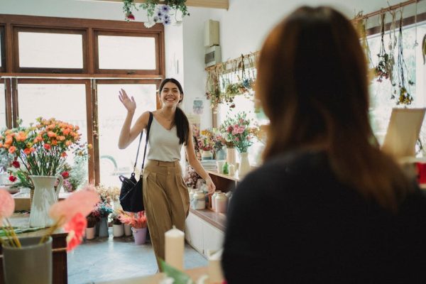 A smiling woman enters a vibrant flower shop, waving to the florist, on a sunny day.
