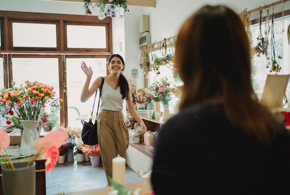 A smiling woman enters a vibrant flower shop, waving to the florist, on a sunny day.