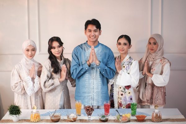 Five individuals in traditional attire celebrating Eid with hands in prayer position over a table with food.