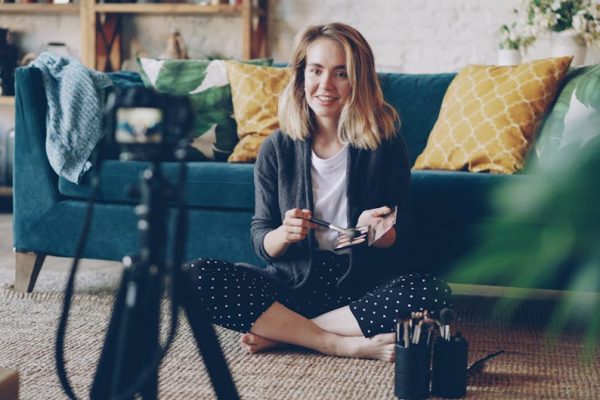 Smiling woman filming a makeup tutorial indoors with brushes and camera setup.