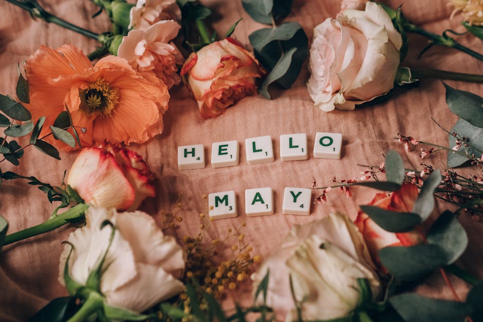 Close-up of scrabble tiles spelling 'HELLO MAY' surrounded by vibrant spring flowers.
