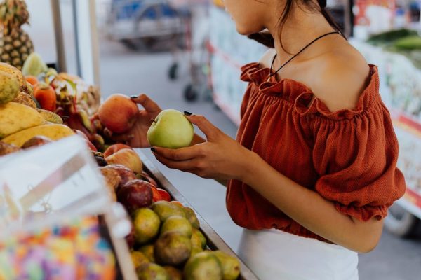 Young woman in orange blouse selecting apples at a bustling outdoor market in daylight.