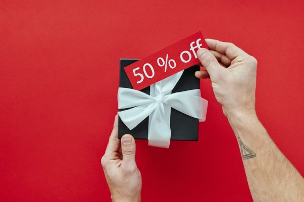 Black gift box with white ribbon held on red background, featuring a 50% off sale tag.
