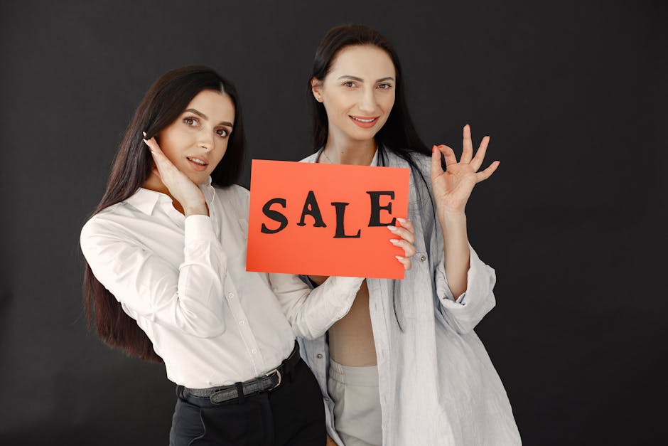Two women in stylish outfits holding a bright sale sign against a black backdrop.