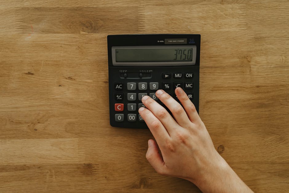 A person's hand pressing keys on a calculator displaying 3750 on a wooden table, top view.