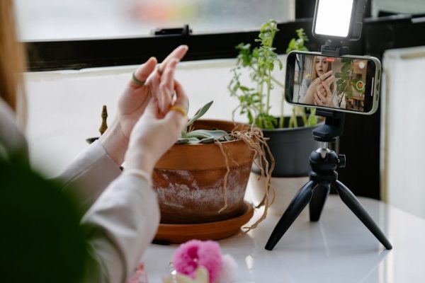 A woman uses a tripod-mounted smartphone to record a video indoors with plants in the background.