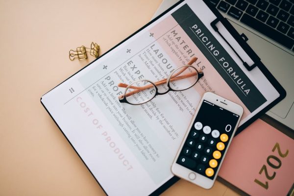 Overhead view of business tools including a phone calculator, pricing formula document, and eyeglasses on a desk.