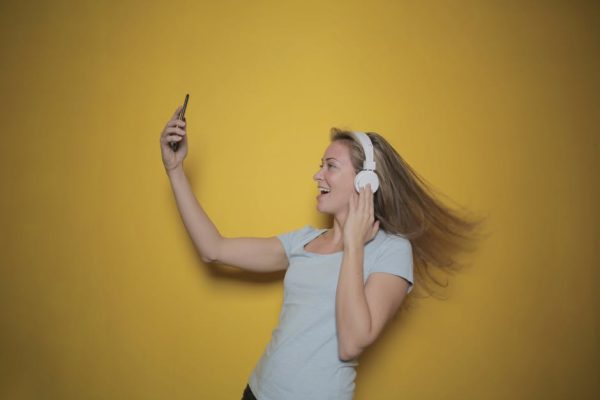 Happy woman wearing headphones taking a selfie against a vibrant yellow background.