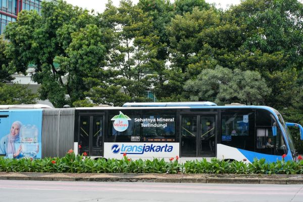 TransJakarta bus passing through a green urban park area with advertisement