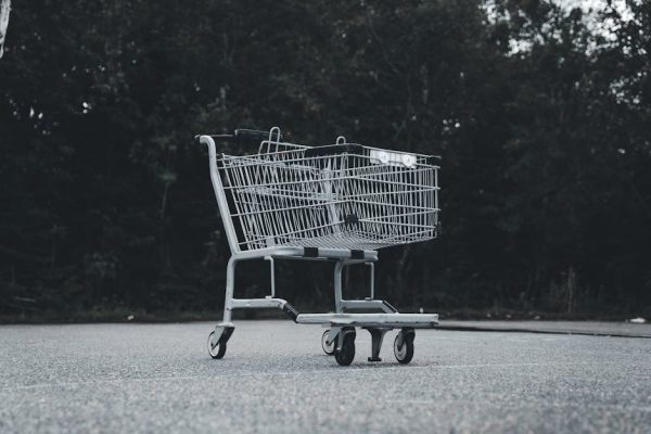 A lone shopping cart sits abandoned on a deserted street with a dark forest backdrop.
