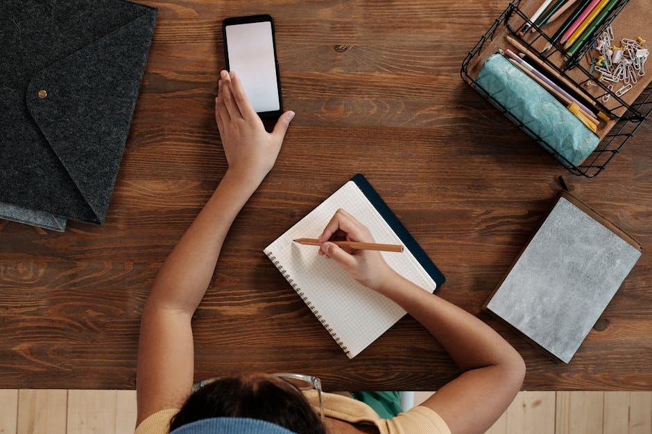 Overhead view of a child writing in a notebook while using a smartphone at a wooden desk.