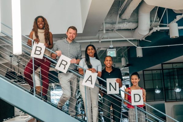 Diverse group of adults smiling and holding 'BRAND' signs on an office staircase.
