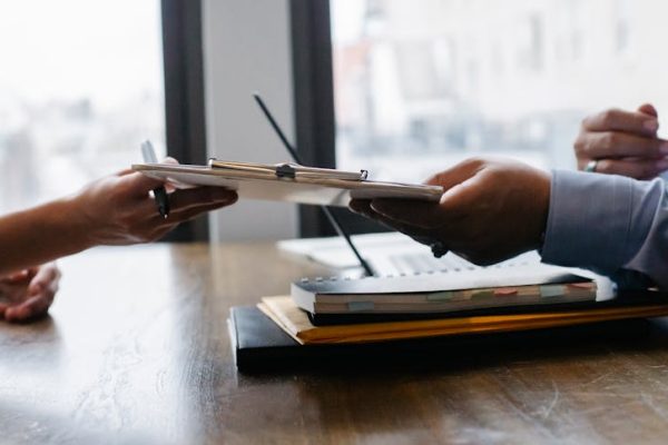 Crop anonymous ethnic woman passing clipboard to office worker with laptop during job interview