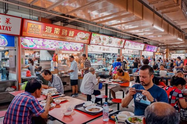 A bustling hawker center with diverse diners enjoying Hainanese chicken rice and various dishes.