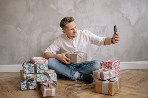 A man sitting on the floor surrounded by wrapped presents, taking a selfie with his smartphone.