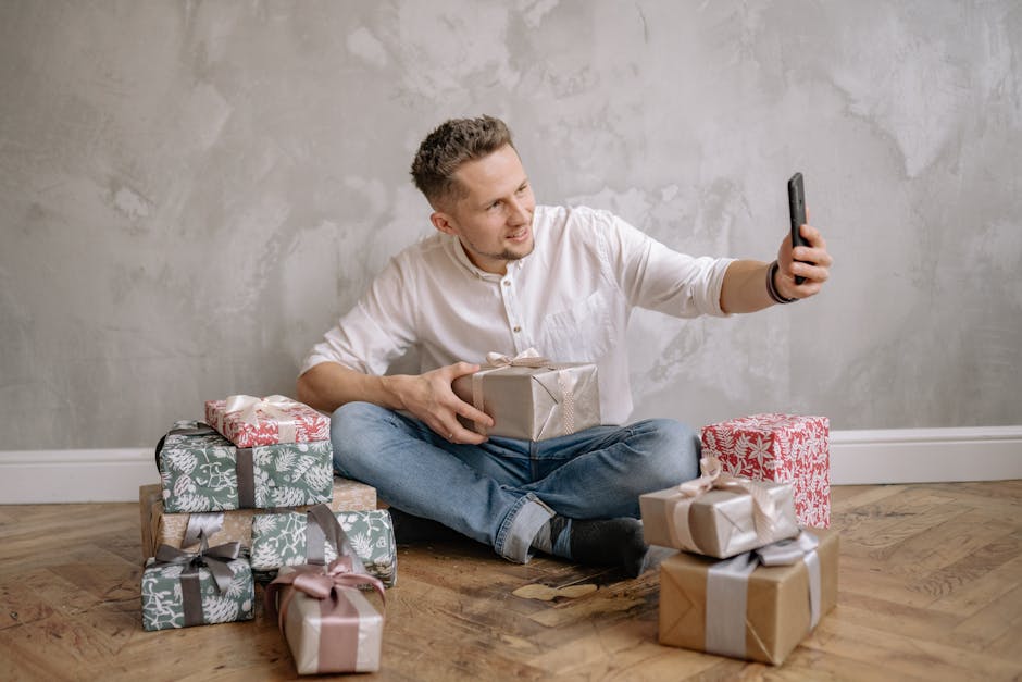 A man sitting on the floor surrounded by wrapped presents, taking a selfie with his smartphone.