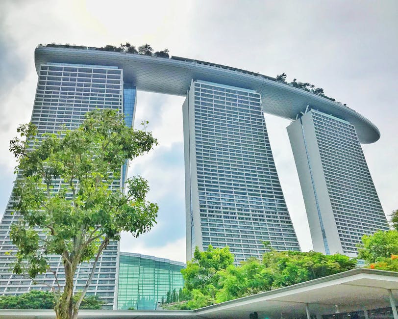 From below exterior of modern stylish skyscrapers complex surrounded with lush green trees against cloudy sky in Singapore