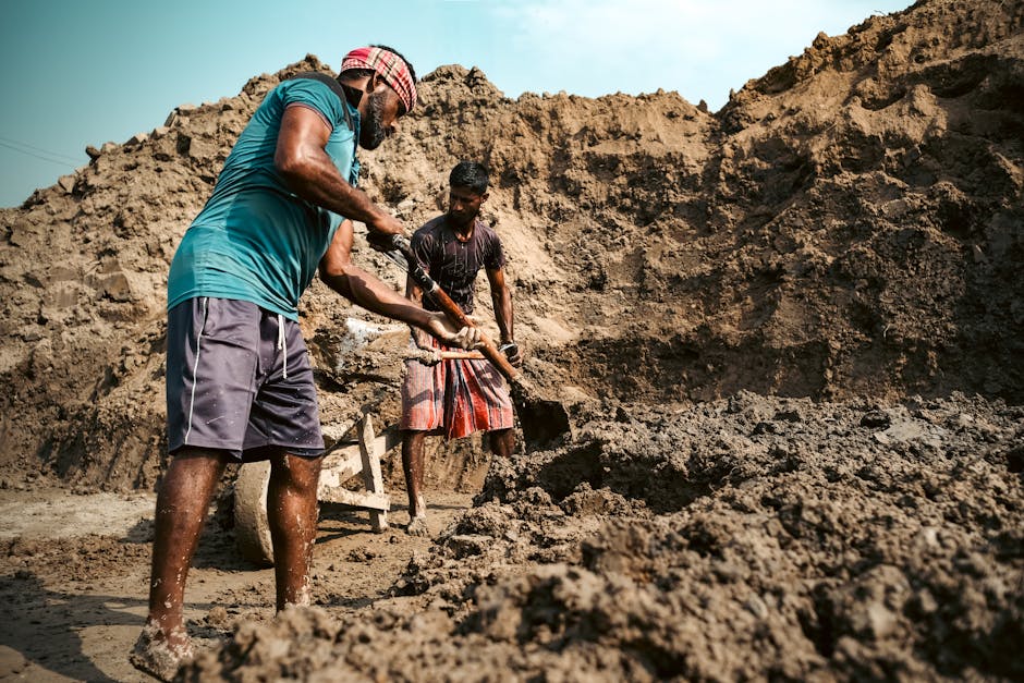 Two men labor in mud at a construction site under a clear sky, showcasing teamwork and hard work.