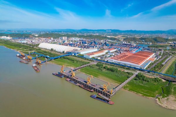 A spacious industrial port with yellow cranes, warehouses, and river access under a clear blue sky.