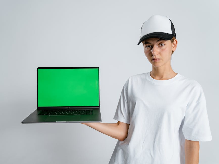 Portrait of a person in a cap holding a laptop with a green screen against a neutral backdrop.