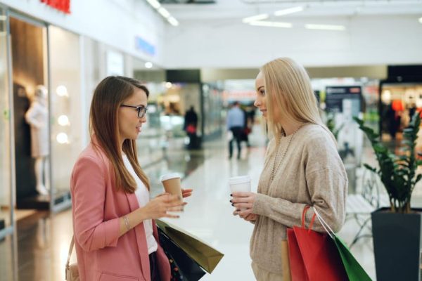 Two women conversing in a shopping mall holding coffee and shopping bags.