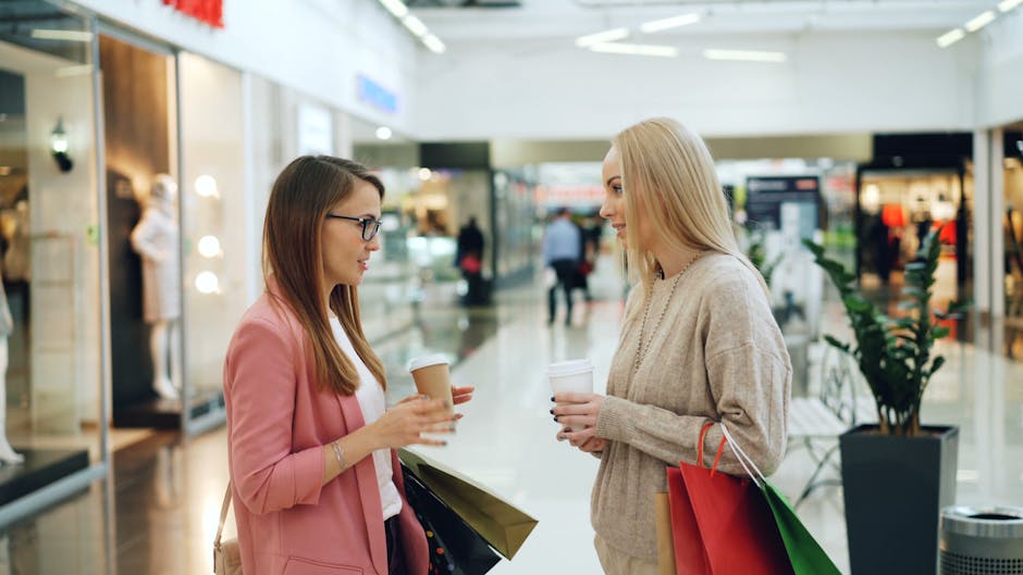 Two women conversing in a shopping mall holding coffee and shopping bags.
