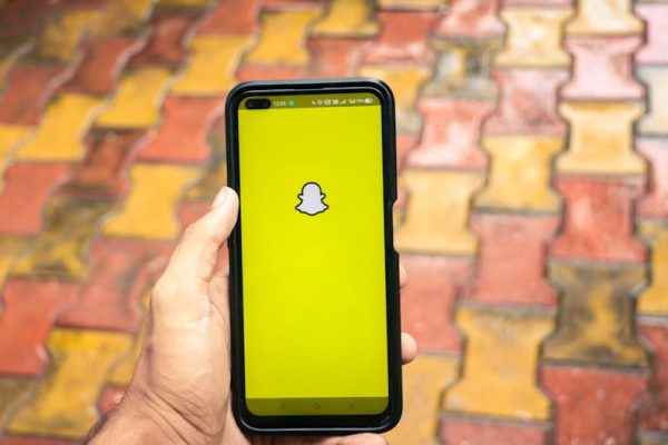 A person holds a smartphone displaying the Snapchat logo against a colorful brick background.