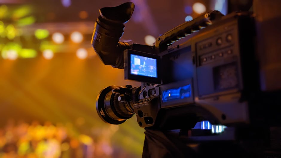 Detailed view of a video camera capturing a live event on stage with blurred lights in the background.